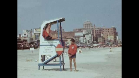 1940s: Lifeguard sits in high chair and another lifeguard stands on the beach, Atlantic City in background. Woman lays on the beach. People walk along the beach. Zeppelin flies from right to left.