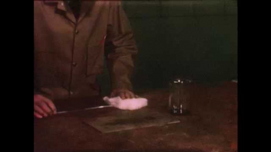 1940s: Man reads thermometer, removes it from inside fabric. Man uses forceps to move fabric to plate and fluff it up. Flames burst from fabric.