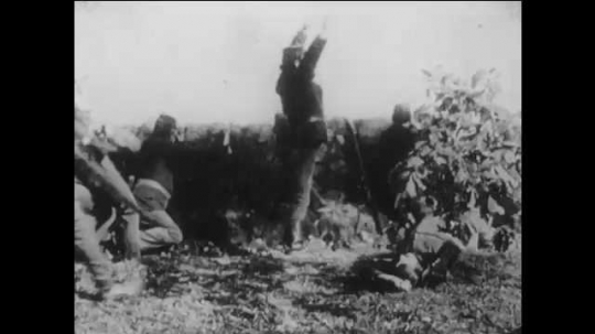 1910s: man in uniform drops dead at wall near shooters during battle. soldier runs from bushes to house and enters door. man enters living room, crouches on floor and hides head as woman laughs.