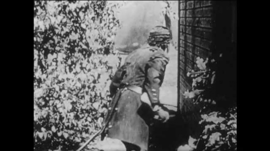1910s: man falls down after explosion on yard. woman looks out window and cries. title card about wounded general. men carry soldier on stretcher into house. men place officer on sofa in living room.