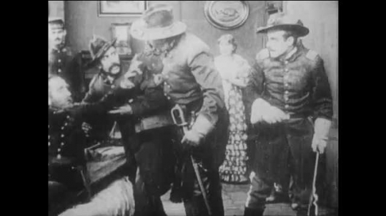 1910s: hurt man sits up, points, orders troops and faints on sofa in living room. soldiers gather behind stone wall. title card about men unite. man in uniform stands up from behind bush near house.
