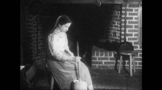 1940s: Sister churns butter by the fire. Boy puts wooden yoke to carry water buckets on shoulders. Mother helps sister at churn. 