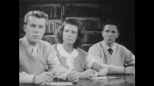 1940s: Students sit at table in library and listen, one shakes his head in disagreement. Student speaks, stands up. One seated student nods head in agreement. Students speak at table. 
