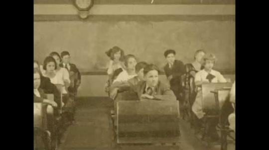 1920s: Students in class nod their heads. Teacher talks to class. Title card appears. Hand writes on paper with pen. Teacher approaches girl at her desk and hands her piece of paper.
