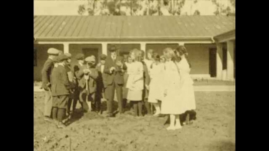 1920s: Students and teacher are gathered around field in front of school. Girl holds tree as they plant it in field. 
