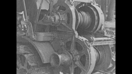 1920s: A cable wind around a reel on a piece of machinery. A drag-line bucket attached to the cable dredges through a river. A man operates the control levers. The bucket pulls earth to the surface. 