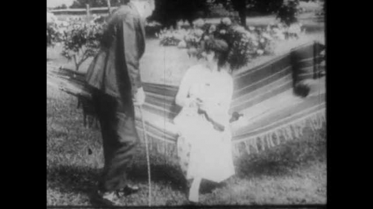 1910s: A man with a top hat chats up a woman sitting in a hammock. Another man looking on pours out a bucket and throws it at him. A third man in a boater hat approaches the woman. They try to sit.