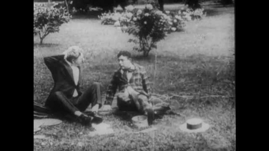 1910s: Two men sit in the grass, arguing. Intertitle reads, 