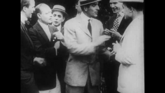 1910s: A group of men at an outdoor party talk amongst themselves. Two men fight. One man pulls the other