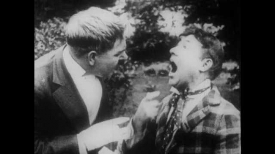 1910s: Two men fight at a picnic. A crowd of men break up the fight. Intertitle reads, 