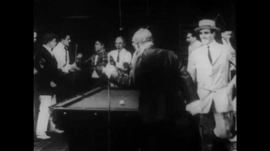 1910s: A crowd at a pool hall congratulates a man. Another man takes a shot, breaking the rack, but the balls stop in the middle of the table. A man tries to put the balls on a shelf, they roll off.