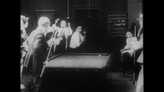 1910s: A crowd congratulates a man for a good shot at a pool hall. The man and his competitor fight and hit each other with pool cues. The crowd laughs.