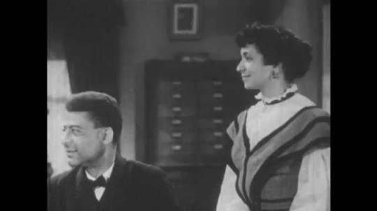 1940s: Booker has a conversation with a woman standing beside his desk.
