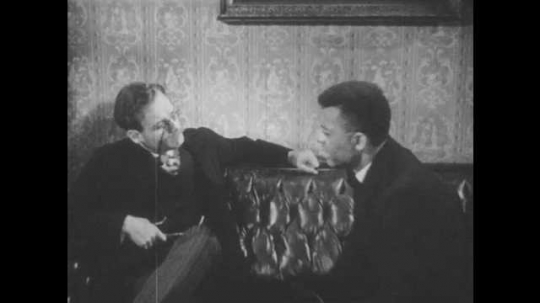 1940s: Booker gestures with his hands as he talks to man wearing pince-nez eyeglasses. Man stands up and checks his pocket watch.