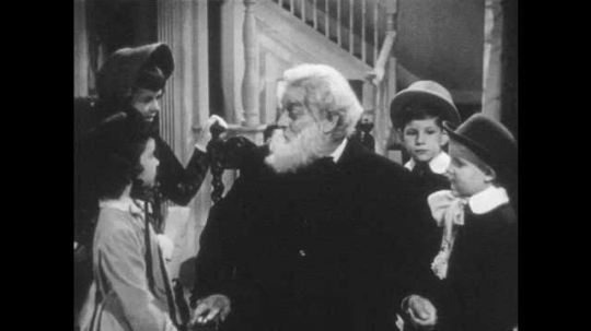 1940s: Old man sits in chair, talks to children. Man places girl onto lap, children gather around man, man talks. 