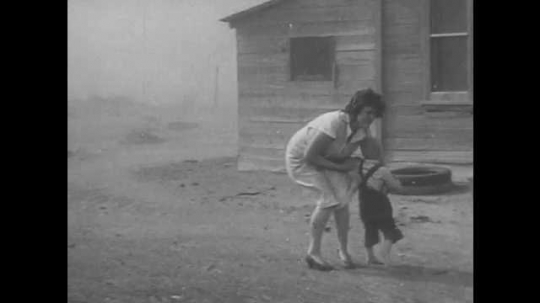 1940s: UNITED STATES: lady picks up baby from yard during dust storm. Vehicles buried under dust in field. Lady runs to house in storm. Man runs to house. Sand piled up by building